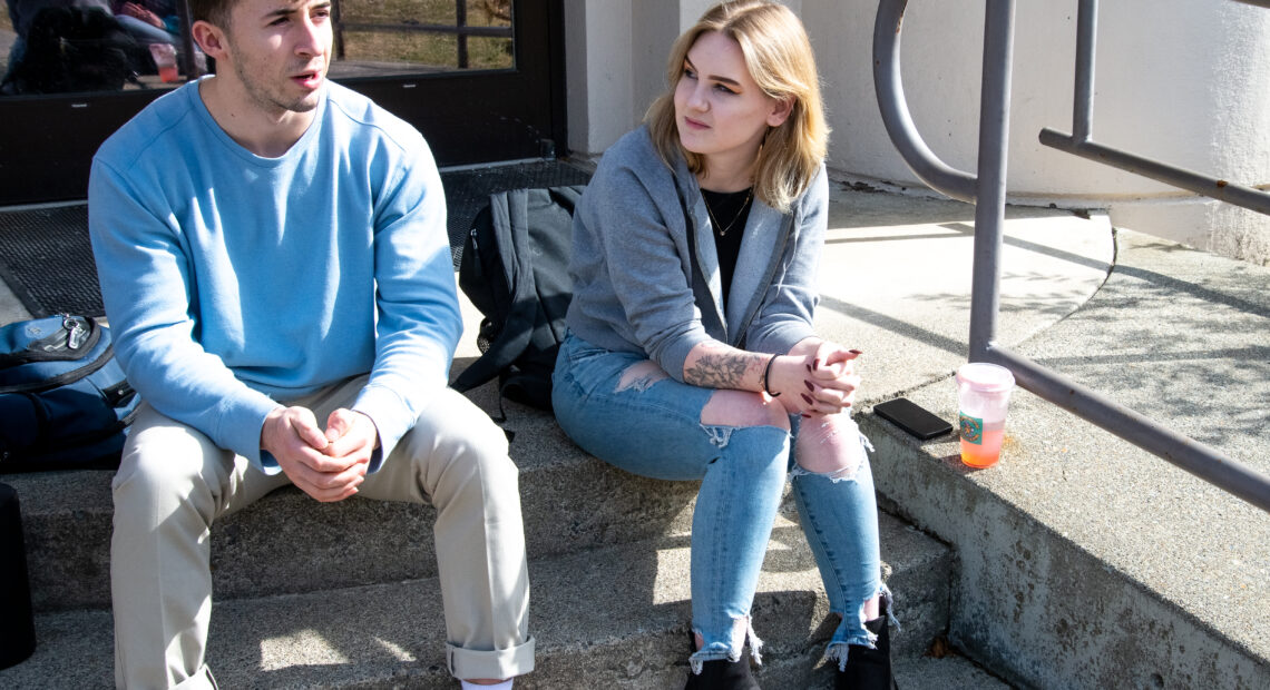 A young man in a blue sweatshirt, khaki pants and brown shoes sits next to a young woman with blonde hair, a grey sweatshirt, jeans and black shoes.