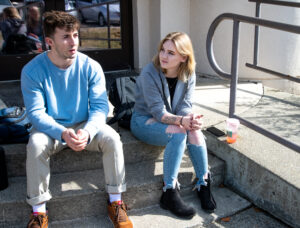 A young man in a blue sweatshirt, khaki pants and brown shoes sits next to a young woman with blonde hair, a grey sweatshirt, jeans and black shoes.