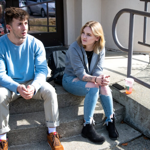 A young man in a blue sweatshirt, khaki pants and brown shoes sits next to a young woman with blonde hair, a grey sweatshirt, jeans and black shoes.