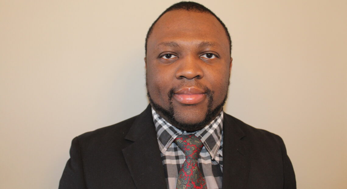 A Black man wearing a suit jacket over grey, plaid button down with a flower-patterned tie looks directly toward the camera.