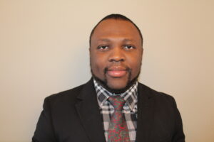 A Black man wearing a suit jacket over grey, plaid button down with a flower-patterned tie looks directly toward the camera.