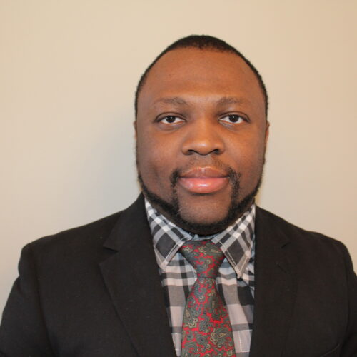 A Black man wearing a suit jacket over grey, plaid button down with a flower-patterned tie looks directly toward the camera.