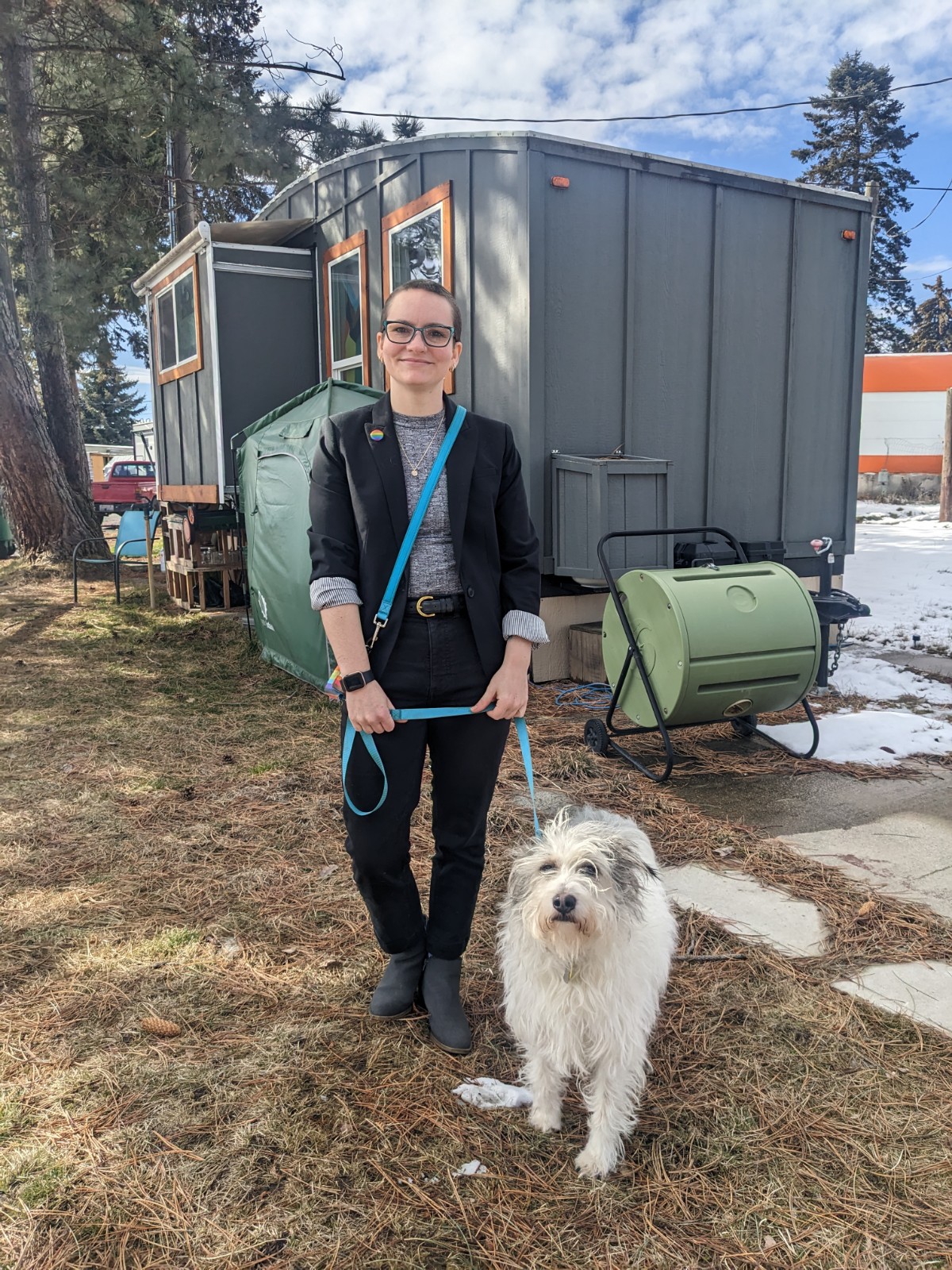 A person with black pants and a black jacket stands in front of a grey tiny home next to a white dog with a blue leash.