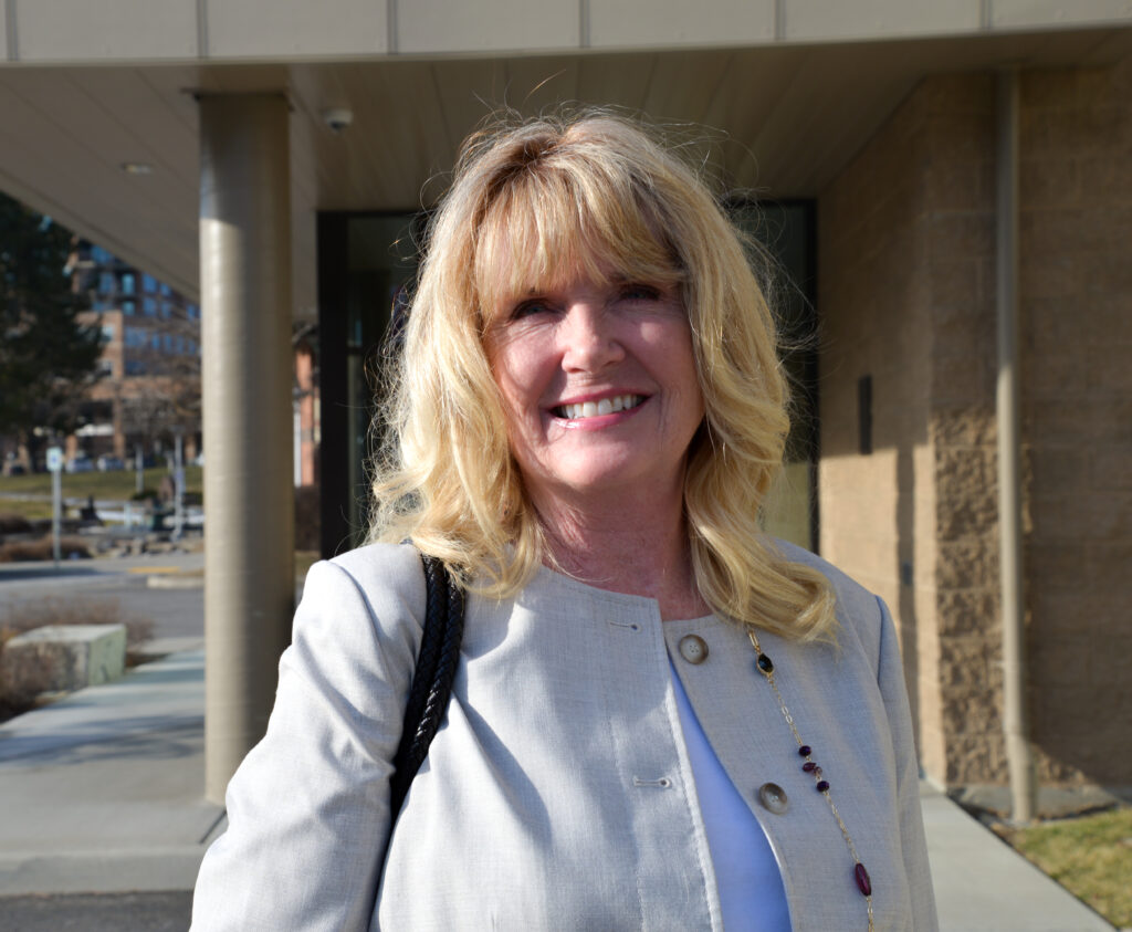 A blonde woman with a tan jacket smiles before Coeur d'Alene City Hall. 