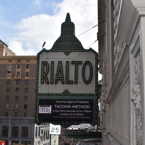 A photo of the marquee outside the Tacoma Opera's premiere of the Tacoma Method opera shows a green rimmed marquee with the word "Rialto" in large block letters. There are office buildings in the background and the signs hangs above a street. In the far background is a blue sky with bright, white clouds.