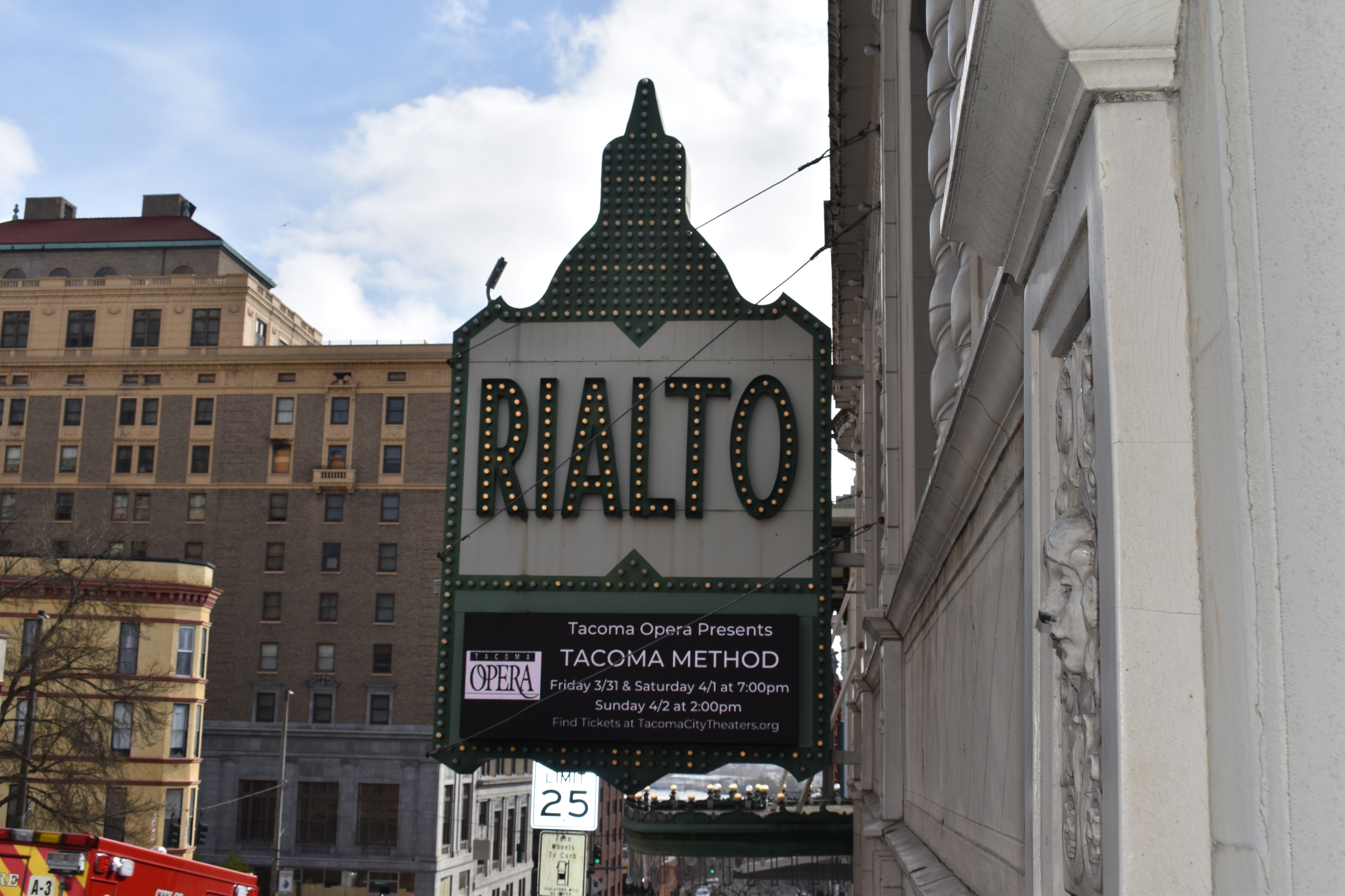 A photo of the marquee outside the Tacoma Opera's premiere of the Tacoma Method opera shows a green rimmed marquee with the word "Rialto" in large block letters. There are office buildings in the background and the signs hangs above a street. In the far background is a blue sky with bright, white clouds.