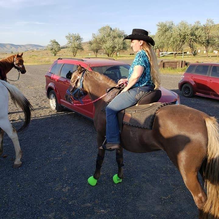 A woman sits on a brown horse. She wears a bright blue t-shirt, a black cowboy hat and blue jeans. The horse is behind a white horse who is mostly out of frame and another brown horse. Two red vehicles are parked in the parking lot where they stand. In the distance is a grassy hill with green trees beneath a light, blue sky.