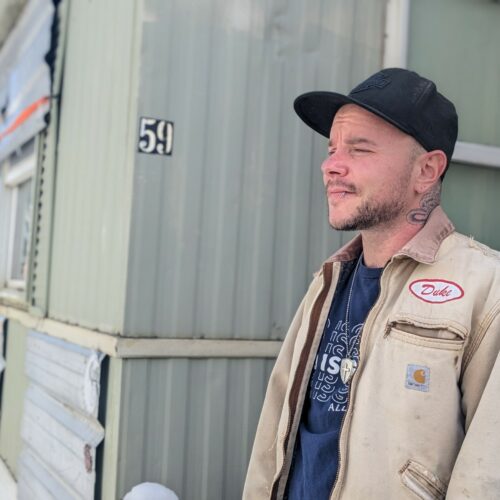 A man with a neck tattoo and a beige jacket stands in front of a green manufactured home in Latah County, Idaho.