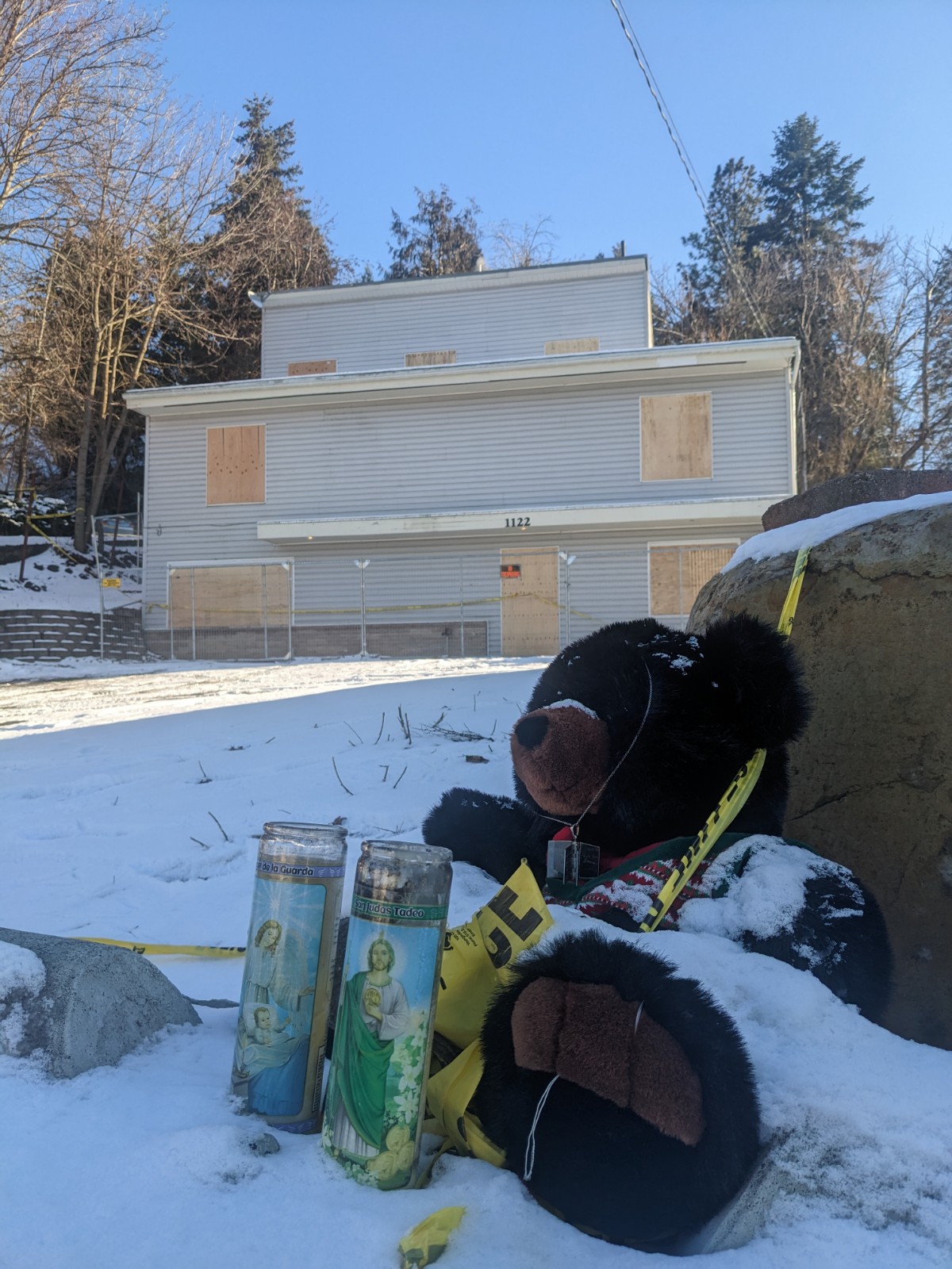 A brown teddy bear and candles sit in snow in front of the white King Road house, with boarded up windows. 