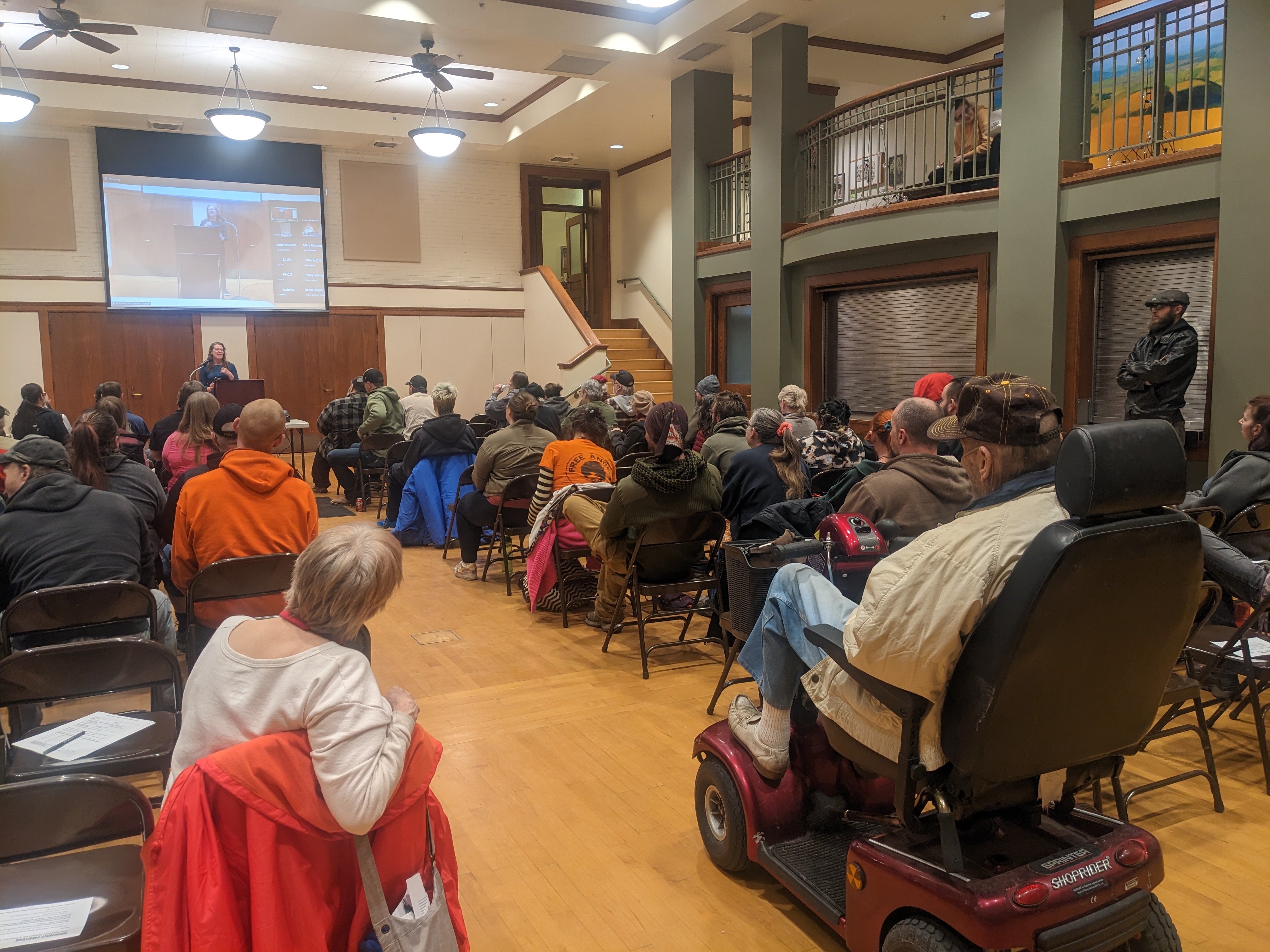 A room with hardwood floors is framed with residents sitting in chairs in behind a projector screen. 