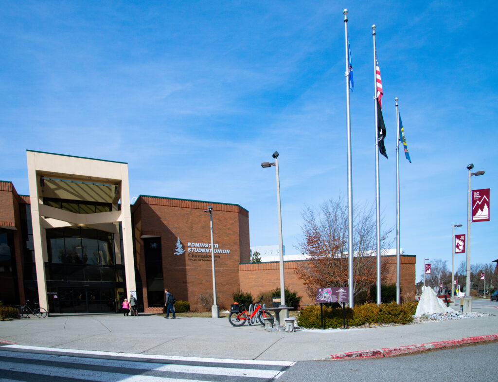 A brick covered student union building sits on concrete with flags flying nearby against a blue sky.