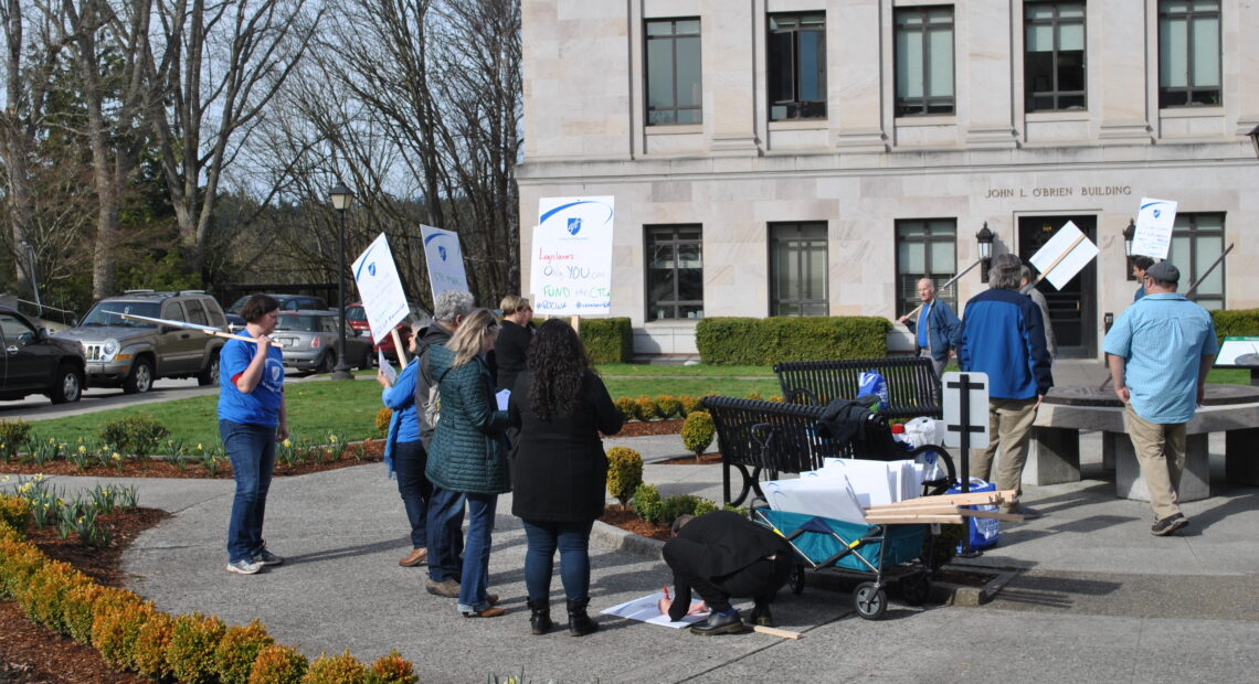 People wearing coats and jeans are gathered in front of a stone building holding picket signs. Someone is kneeling over a poster on the ground as they draw out a message.