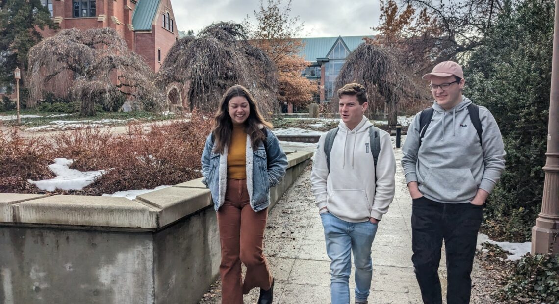 Three students with backpacks walk on pavement lightly covered in snow in front of a brick building with a green roof.