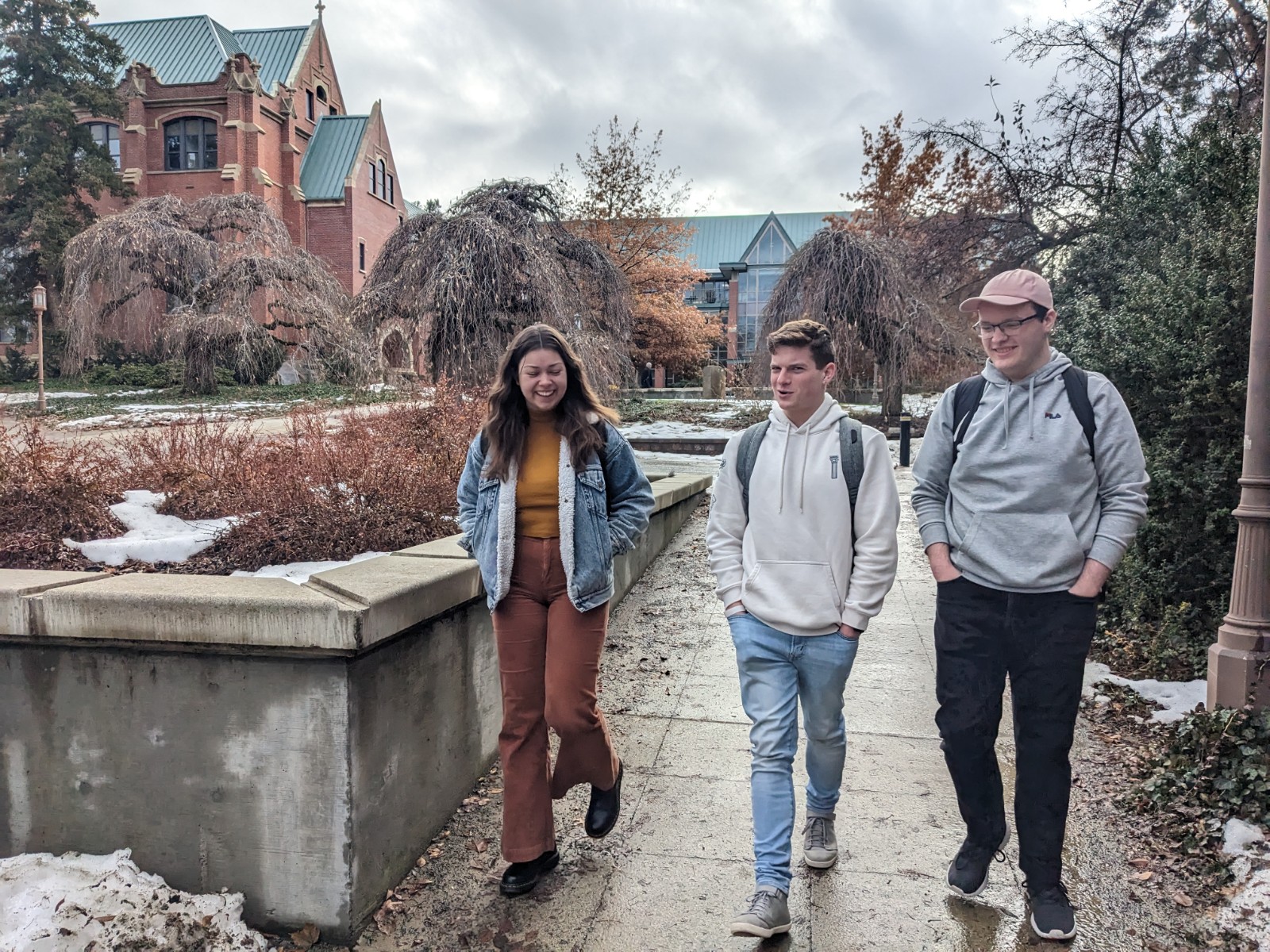 Three students with backpacks walk on pavement lightly covered in snow in front of a brick building with a green roof.