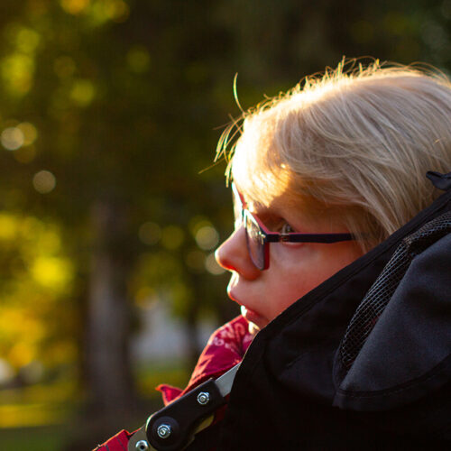A woman with short, blond hair sits facing toward the left in profile of the camera and looking through dark-rimmed glasses. In the background are green trees out of focus in golden sunlight.