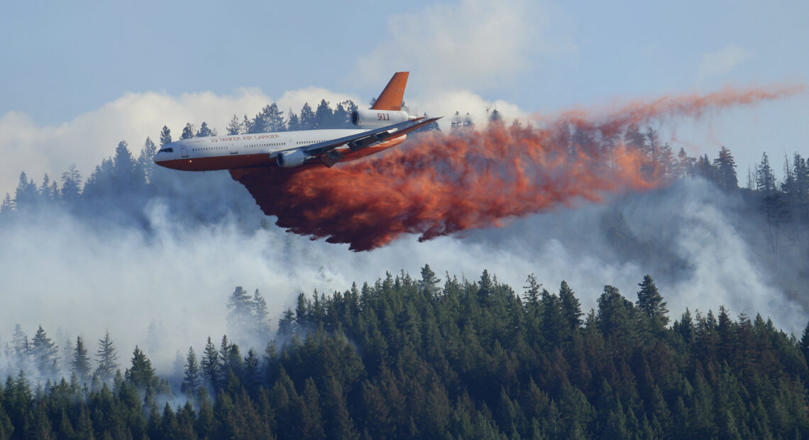 A white tanker plane drops bright orange flame retardant onto a smokey forest of evergreen trees.