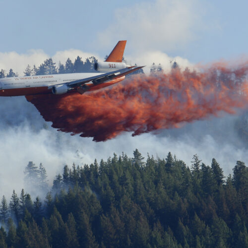A white tanker plane drops bright orange flame retardant onto a smokey forest of evergreen trees.