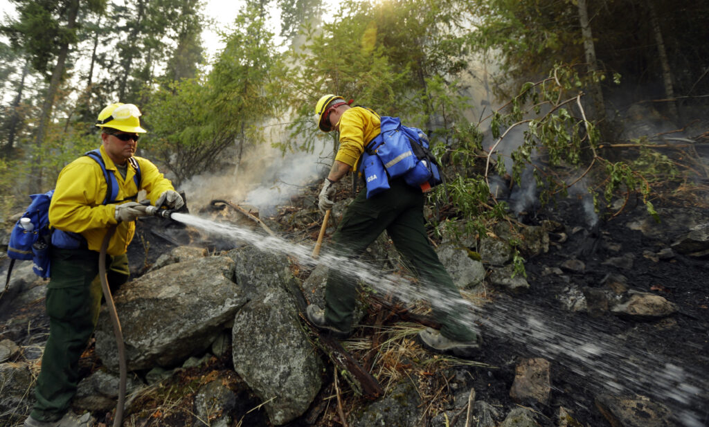 Two national guard firefighters with yellow jackets and yellow hardhats spray down a smokey and rock covered hillside surrounded by green trees. 