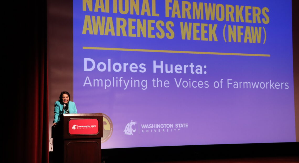 Dolores Huerta speaks at a podium at Washington State University. She wears a black shirt and a turquoise jacket.