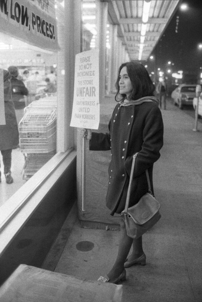  Dolores Huerta is protesting and standing in front of a window of a Safeway in Seattle, Washington. She is holding up a sign mounted on a stick in front of her with her right hand. Her left hand rests in her pocket with a purse looped around her arm. Her sign reads: "Please do not patronize. This store unfair. Grape workers united farm workers." 