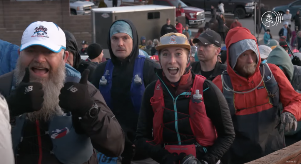 Runners wearing water bottle backpacks, hats, headbands, headphones, and long sleeves walk toward finish line and smile toward the camera.