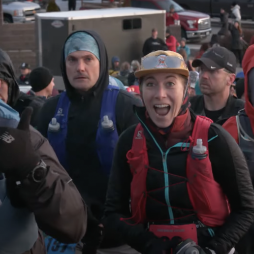 Runners wearing water bottle backpacks, hats, headbands, headphones, and long sleeves walk toward finish line and smile toward the camera.