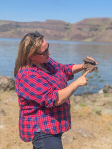 A women in a blue and red plaid shirt stands in front of a blue river. She is holding two gray rocks on top of each other. in the background, on the other side of the river, there are rocky hills and a blue sky.