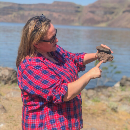 A women in a blue and red plaid shirt stands in front of a blue river. She is holding two gray rocks on top of each other. in the background, on the other side of the river, there are rocky hills and a blue sky.