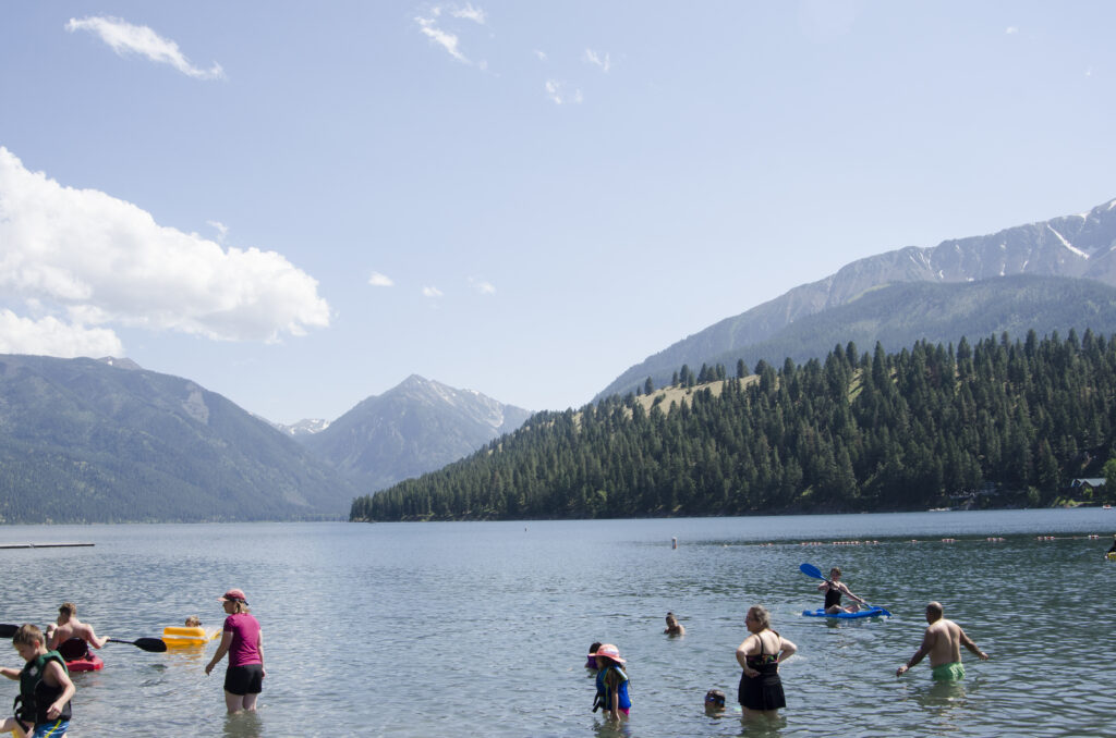 People of all ages stand in blue lake waters against an evergreen covered hilltop under a cloudy blue sky. 