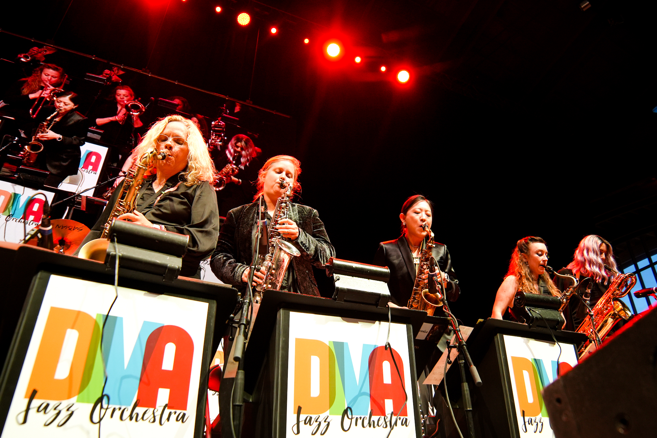The DIVA Jazz Orchestra performers stand under red lights behind podiums with the bright yellow, green, blue and red title letters of their group emblazoned on the front. The lead performers are a group of women all wearing black tops and playing jazz band instruments.