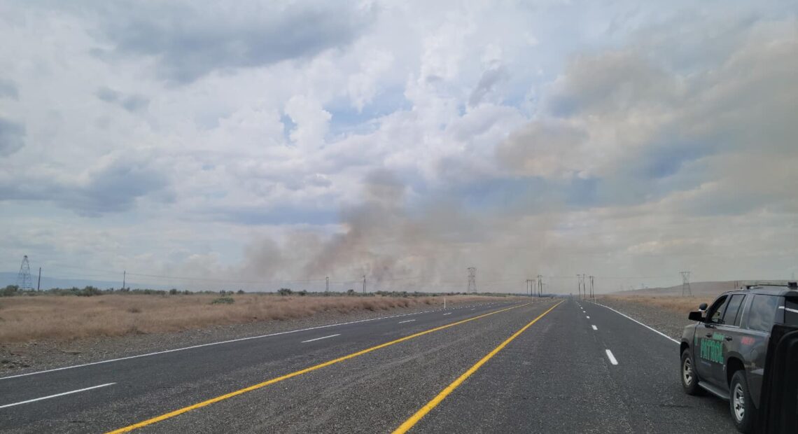 A two-lane road stretches into the distance. It's surrounded by brown dessert landscape on either side. In the background of the picture, a small plume of brown smoke rises into blue, cloudy skies.