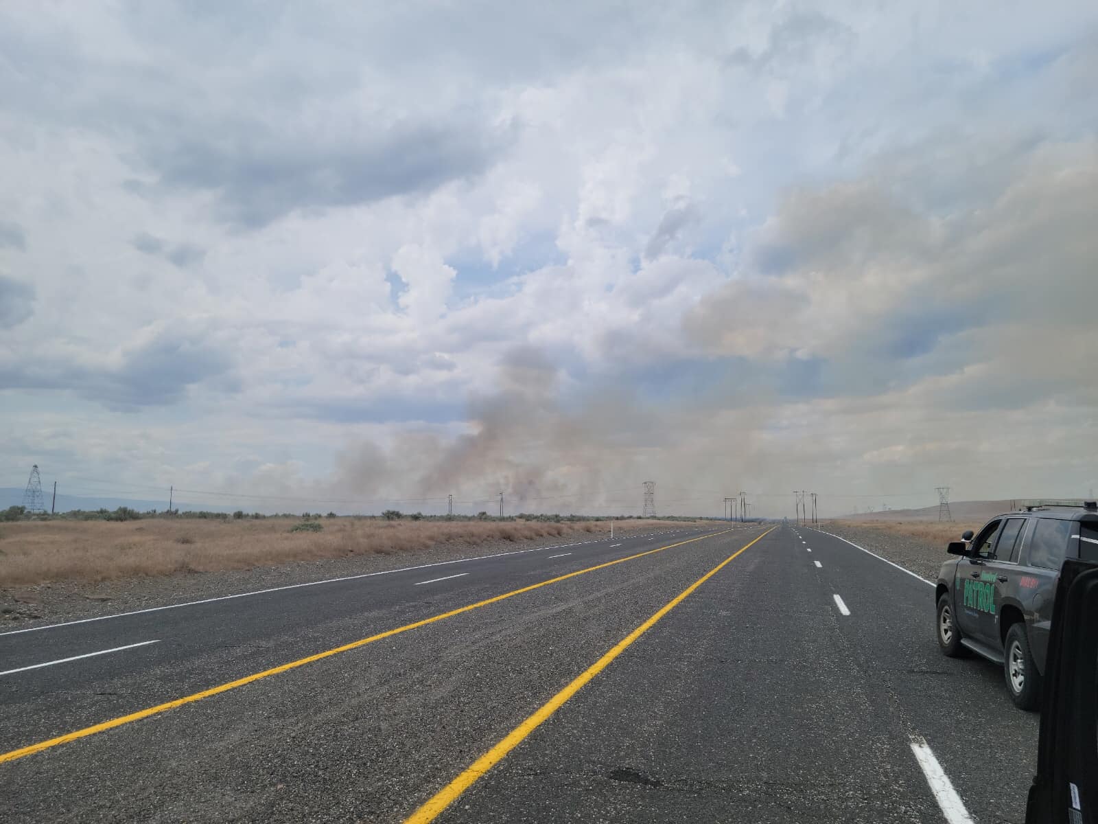 A two-lane road stretches into the distance. It's surrounded by brown dessert landscape on either side. In the background of the picture, a small plume of brown smoke rises into blue, cloudy skies.