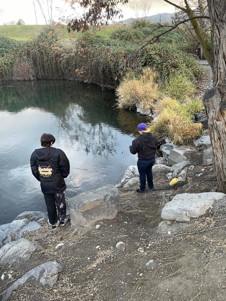 Two students dressed in black stand over a pond surrounded by rocks and green plants.