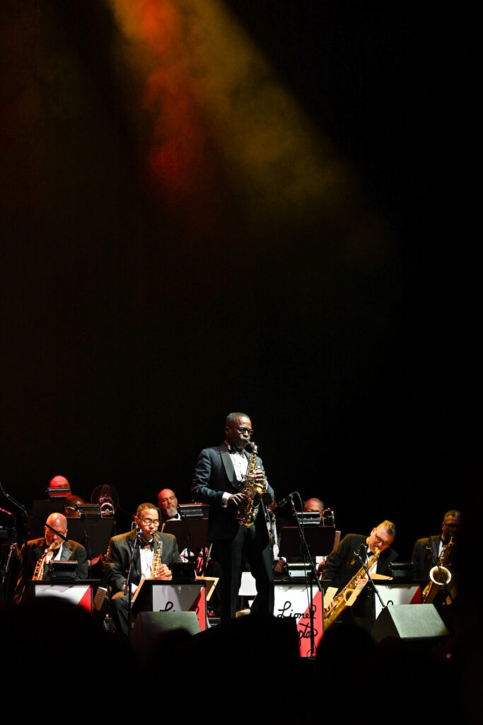 Set against a back black drop, a spotlight illuminates two channels of red and yellow fog above a stage where a big band is performing. Artists sit in rows, playing band instruments, in the front stands a black man in a tuxedo playing a saxophone, closing his eyes as he performs. In the foreground, out of focus and in silhouette, are the dim outline of the crowd watching the performance.