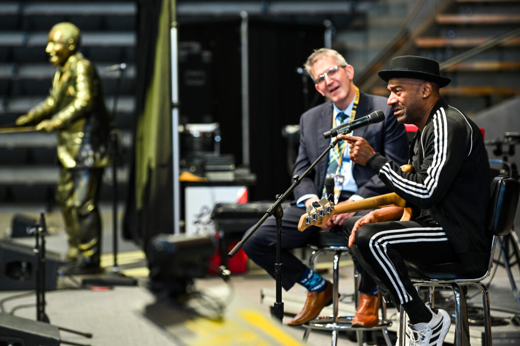 Marcus Miller sits in a metal chair in profile. He wears a black track suit with white stripes and a black hate. He's holding a guitar, with only the neck visible) and pointing toward the audience. In the background, another man looks on as they sit on a stage in an auditorium.