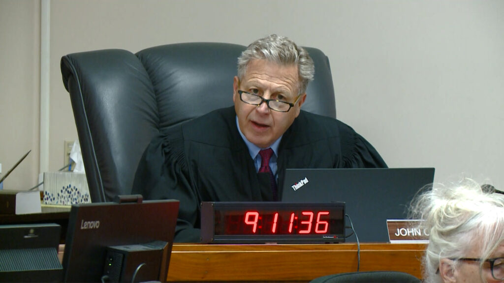 A judge with grey hair and glasses sits at a brown bench in a big black chair. 