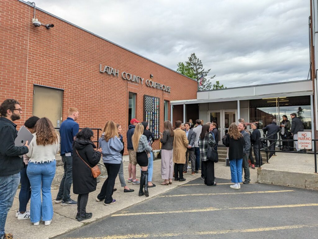 A long line of people stand before a brick building labeled Latah County Courthouse. 