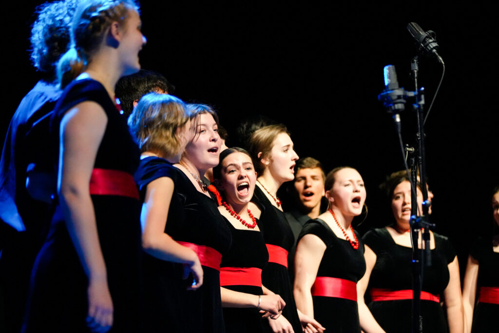 A row of young women stand in a the front row nearest an out-of-focus microphone and captured singing. They wear the same black, short-sleeved velvet dresses with red sashes and matching red necklaces. In the back row, stand a group of young men, mostly obscured by the women but clearly wearing black suits and singing as well.