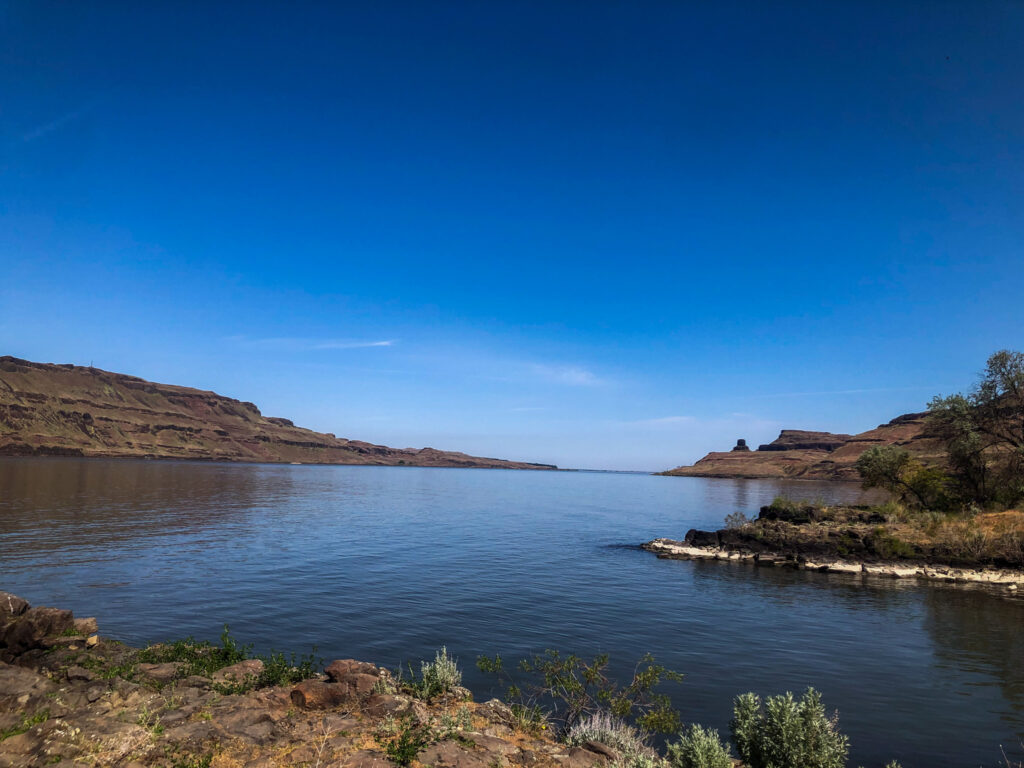 A blue river is surrounded by brown rocky hills. The sky is very blue with a few white, wispy clouds.