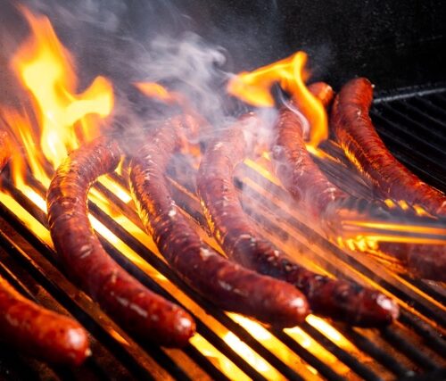 Six brown sausages sit on top of a black grill. Orange flames crawl up the grill and onto the sausages.