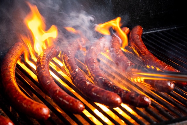 Six brown sausages sit on top of a black grill. Orange flames crawl up the grill and onto the sausages.