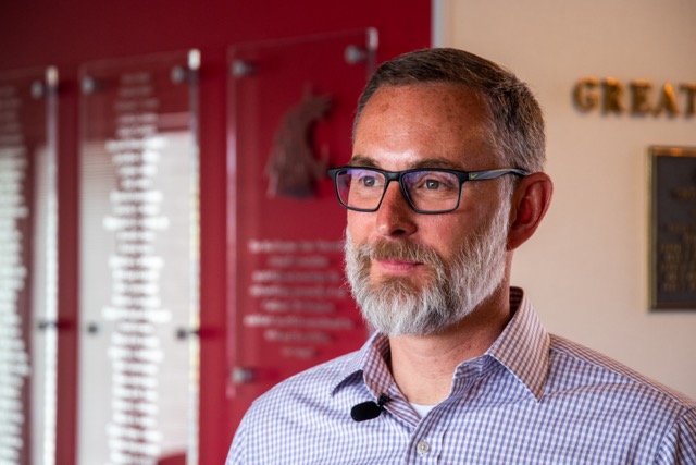 A portrait of a man in black glasses. He has grey hair and a grey and brown beard. He's wearing a blue checked button down shirt. He's standing in front of a crimson, silver and white wall.