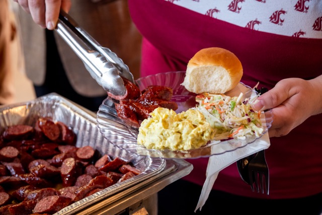 In the front of the picture there is a silver pan with brown sausages in it. In the center of the picture, a person is holding silver tongs with the brown sausages in them. The person is also holding a clear plate with sausages, bread, coleslaw and potato salad. The person is wearing a maroon and white cougar and WSU shirt. The person's head isn't in the photo.