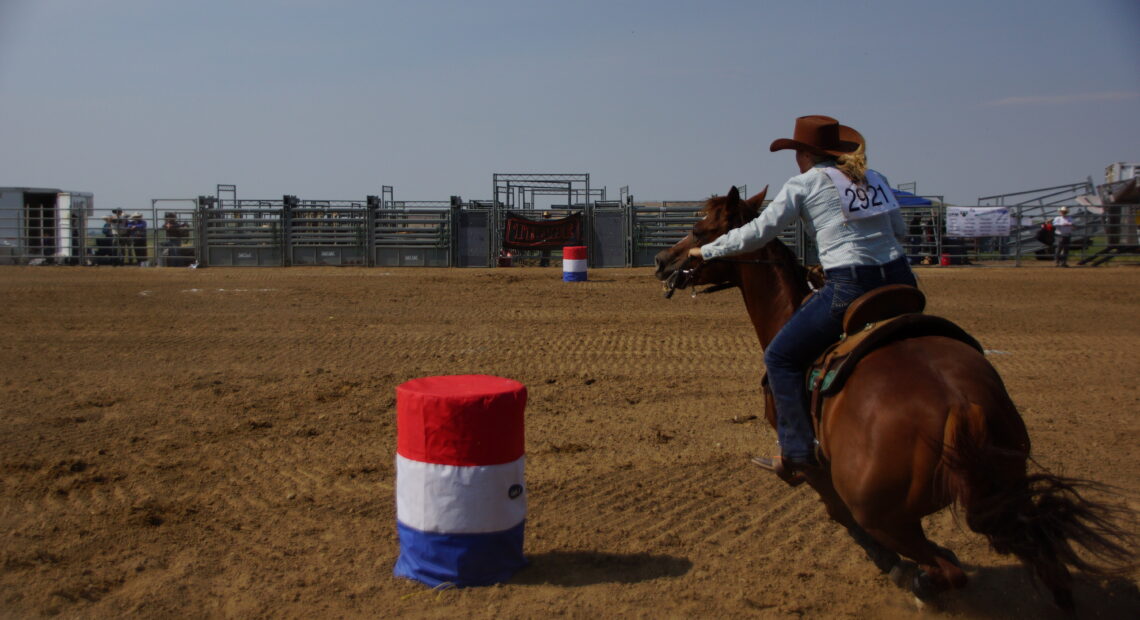 A woman with a blue shirt and a cowboy hat barrel races atop a brown horse at the Denver Gay Rodeo 2021.