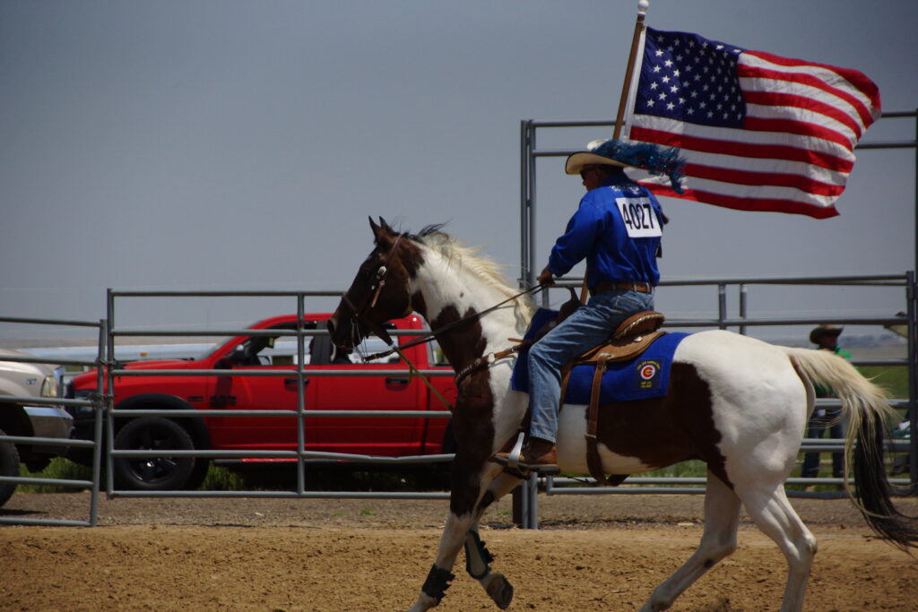 A man in a blue shirt and blue jeans rides a white and brown horse while carrying an American flag. 