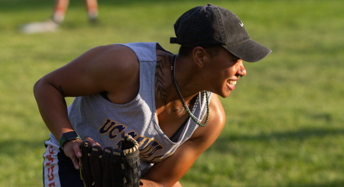 A young baseball player wearing a black ball cap grins toward the right of frame. They wear two necklaces, one silver metal and one green bead, and a botanical tattoo of leaves encircle their neck. They crouch, with a baseball mitt in one hand. In the background a field of blurred out grass is visible.