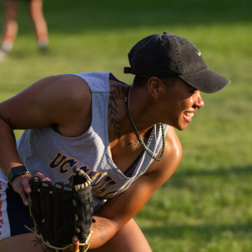 A young baseball player wearing a black ball cap grins toward the right of frame. They wear two necklaces, one silver metal and one green bead, and a botanical tattoo of leaves encircle their neck. They crouch, with a baseball mitt in one hand. In the background a field of blurred out grass is visible.