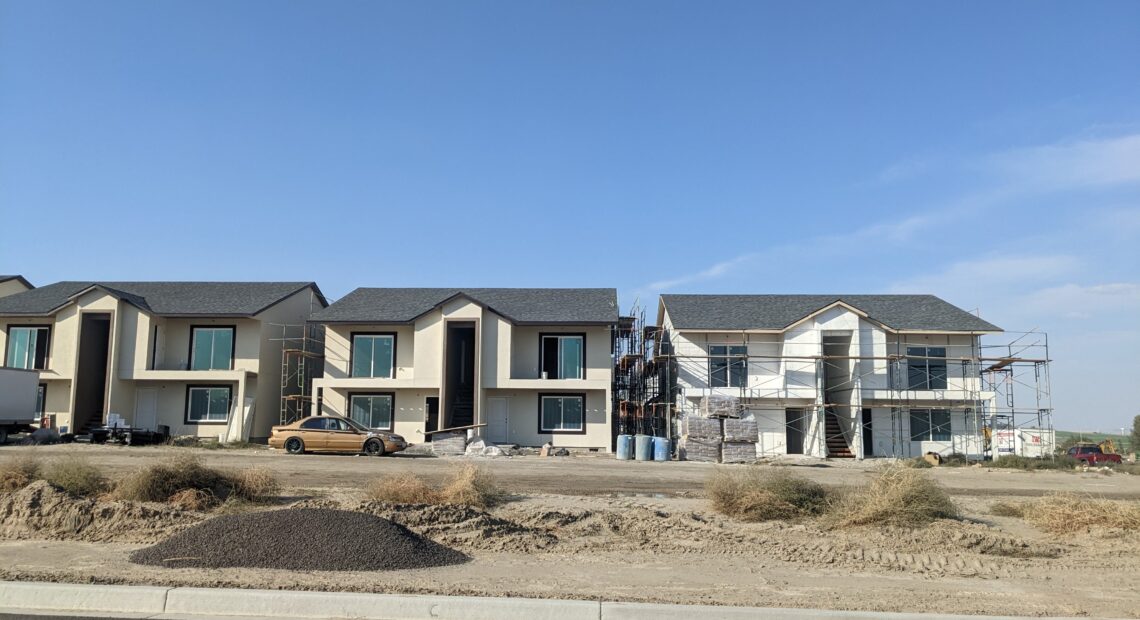 White and grey houses under construction sit on sandy ground under a blue sky.