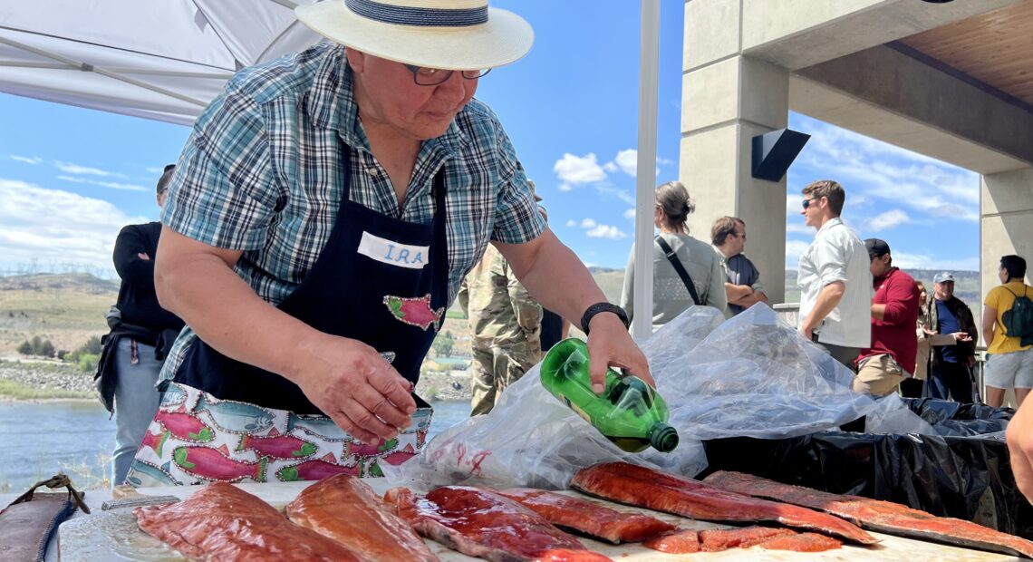 A man in a blue plaid shirt, black apron and white fedora holds a green bottle of olive oil above six pink filets of salmon. The filets are sitting on a white plastic table. A crowd of people stands behind him looking at a blue river.