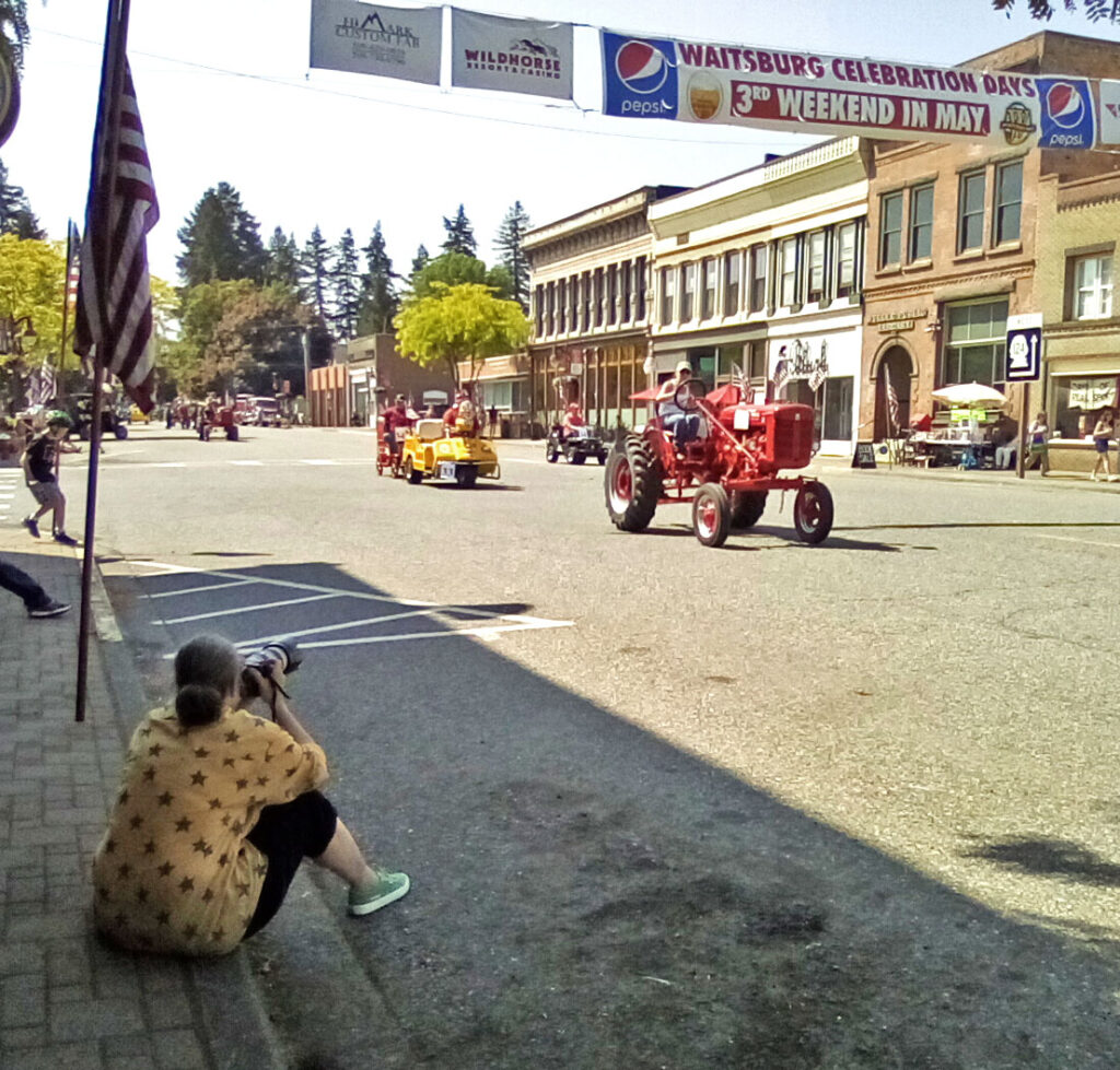 A woman in a yellow shirt with maroon stars on it sits on a grey sidewalk. She is holding a grey and white camera. In the middle of the street, a red tractor and a yellow car are driving past. There is an American flag in the left hand corner of the photo. In the background, there are tan and white buildings.